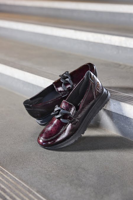 Close-up van bordeauxrode lakleren Riekerdames loafers met zwarte details en gesp, geplaatst op een moderne betonnen vloer