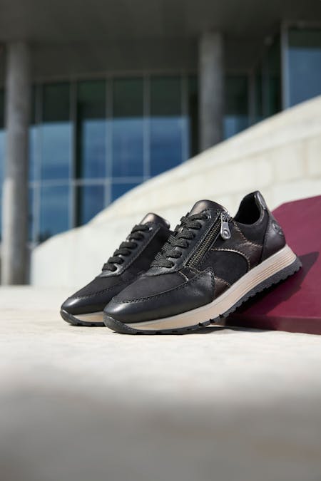 Close-up van zwarte leren dames sneakers met ritsdetail en witte zool, gefotografeerd buiten bij een modern gebouw.